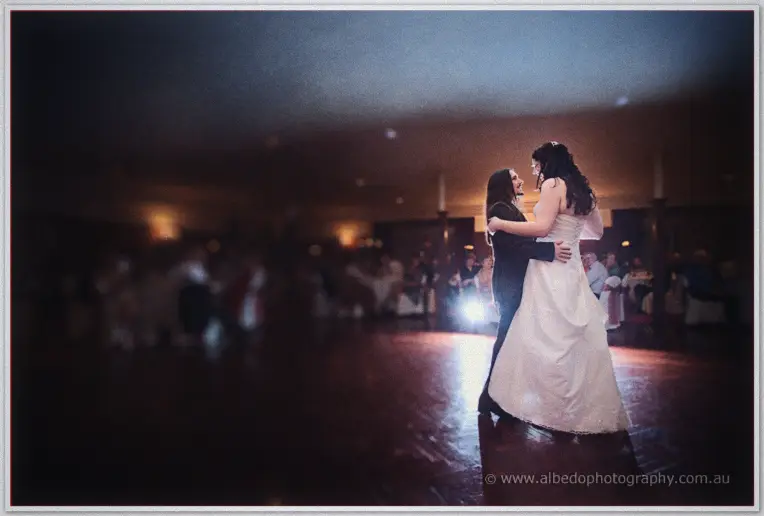 Jenn and Lance share their first dance on the wooden floor as blurred guests watch from dim lamplight