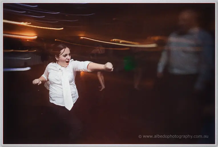 A guest in white shirt and tie dances wildly as streaks of light blur across the dim reception floor