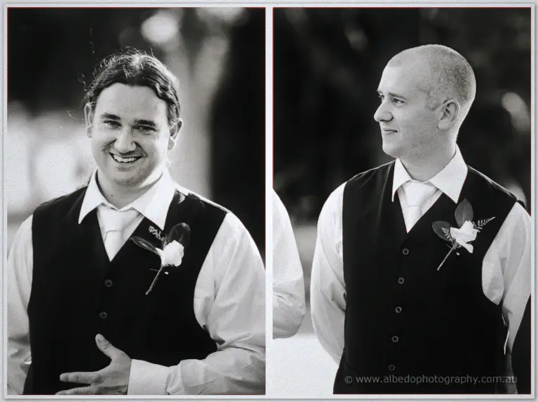 Black and white portraits of two smiling groomsmen in vests with white rose buttonholes by the water