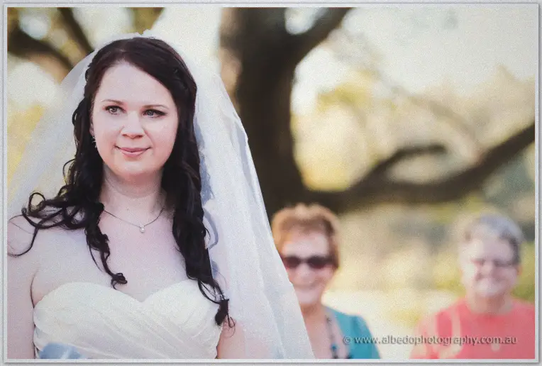 Jenn walks down the aisle beneath her veil, soft light and blurred guests behind her