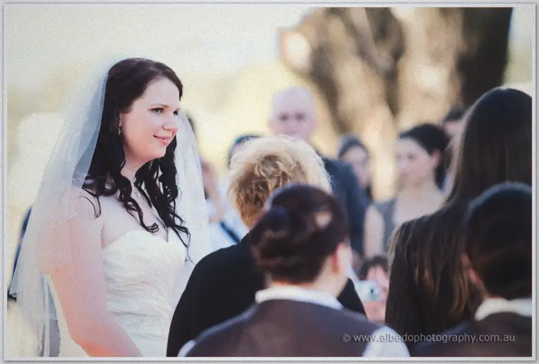 Jenn smiles toward her guests during the outdoor ceremony, heads of the gathered crowd in the foreground