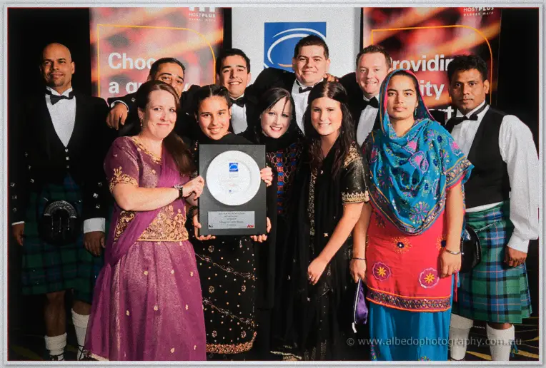 Award winners and staff pose with a plaque on stage, kilts and formal gowns under gala stage lights