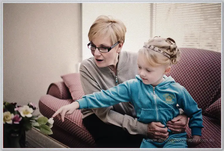 A grandmother steadies the little girl as she reaches for flowers on a lounge-room table