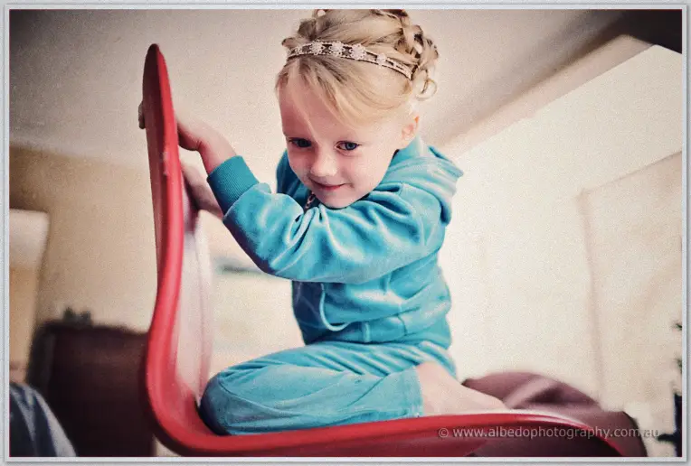 A young flower girl in a turquoise tracksuit and beaded headband perches on a red chair at home