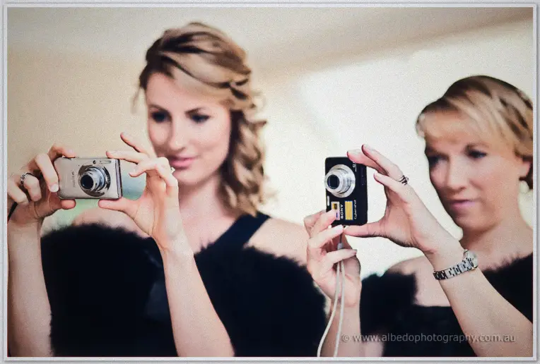 Two bridesmaids in black feathered wraps aim their compact cameras playfully