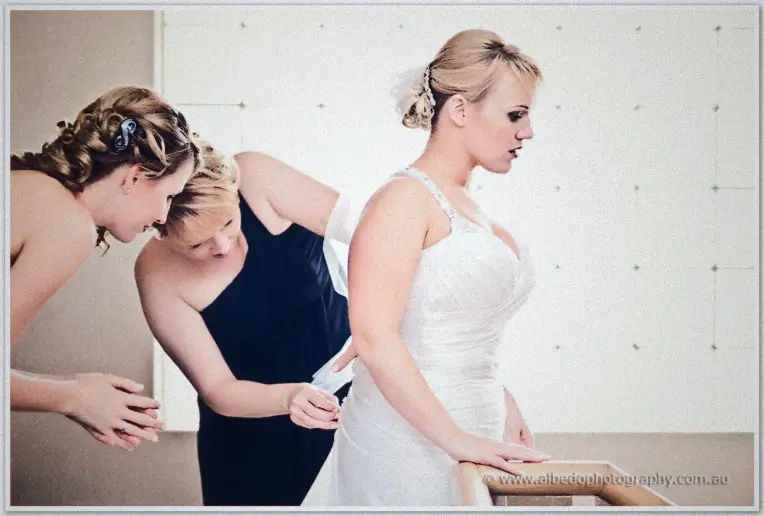 Bridesmaids lace the back of Emily's gown as she leans on a stair rail in bright light