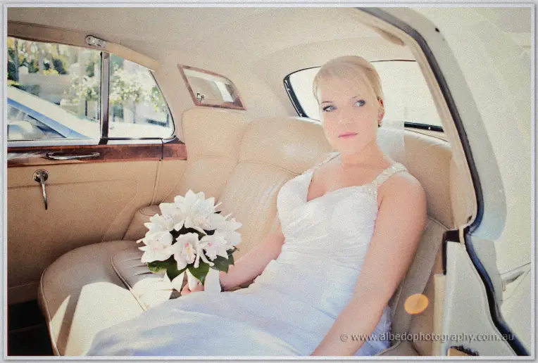 Emily sits in the back of a cream vintage limousine holding her white orchid bouquet in daylight