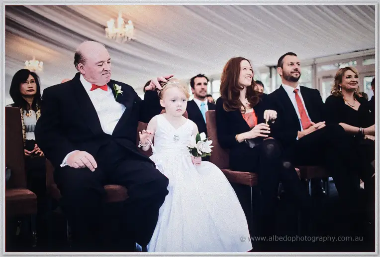 The flower girl sits beside Emily's father as guests watch the marquee ceremony