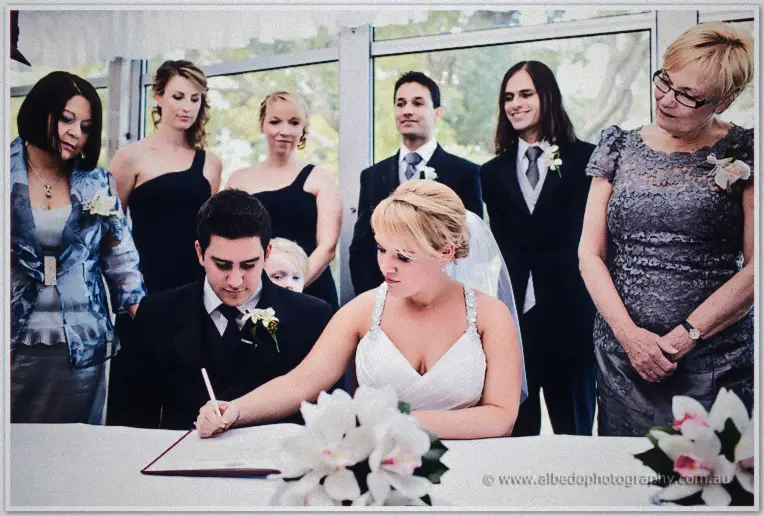 Emily signs the marriage register while family and the bridal party gather round