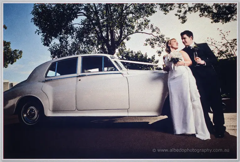 Emily and Quentin toast beside their white vintage car at dusk, ribbon tied to the door