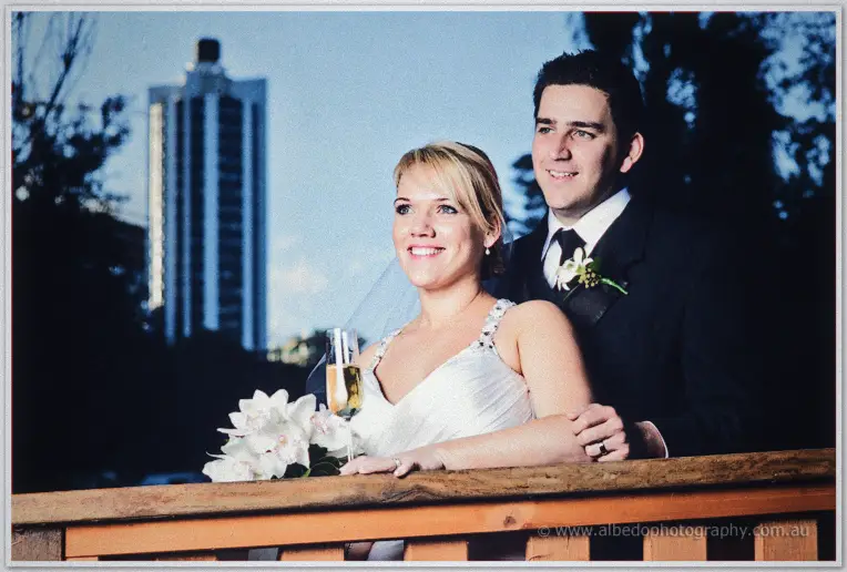 Emily leans on a timber railing with Quentin behind her, champagne in hand, city tower behind