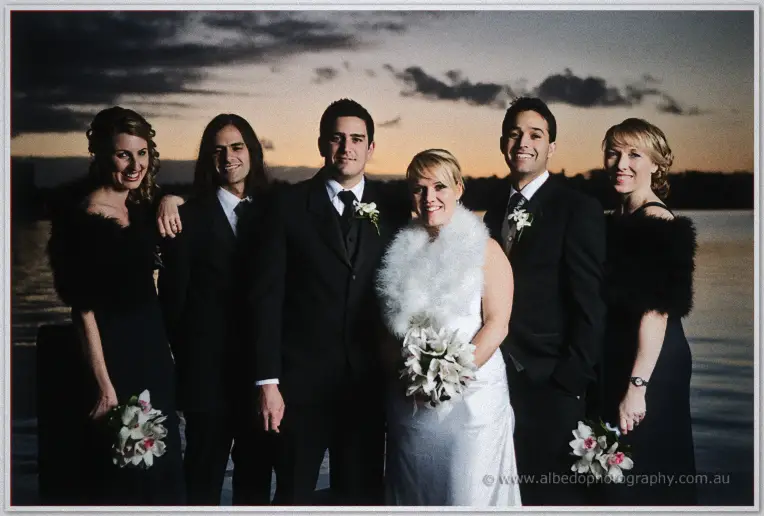 The full bridal party gathers by the river at sunset, Emily in a white feather wrap