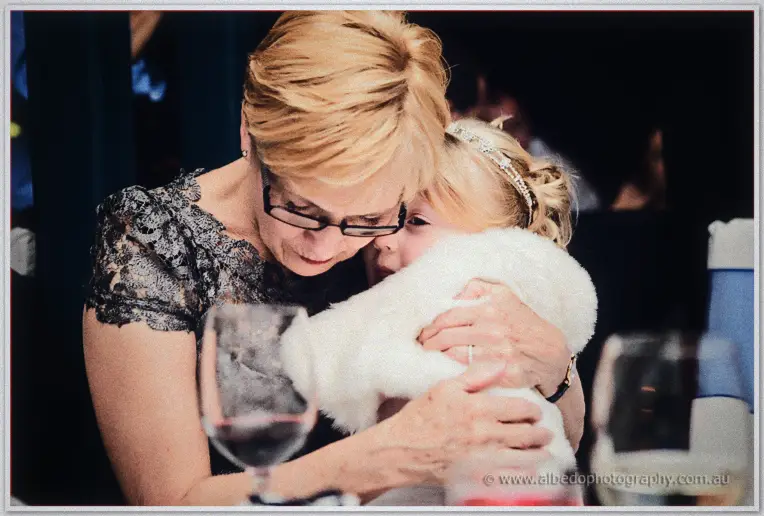 Emily's mother hugs the flower girl close at the reception table amid wine glasses