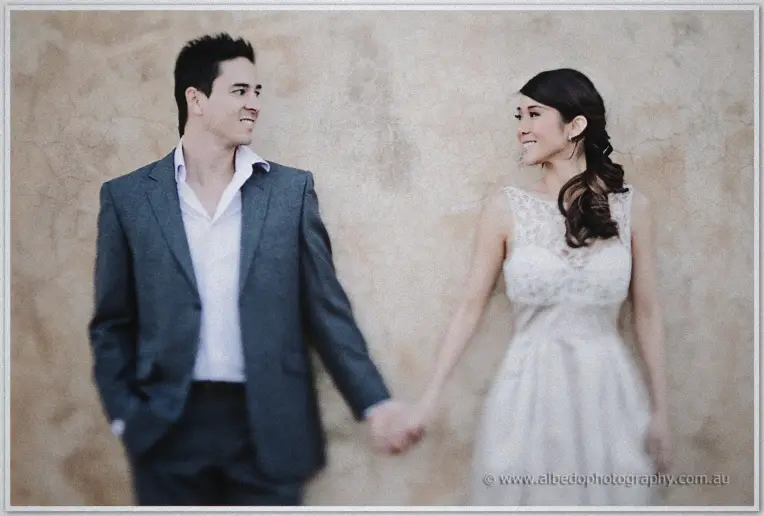 Pre wedding Fremantle | Jade and Aaron Jade and Aaron hold hands and grin at each other before a mottled cream wall in soft light