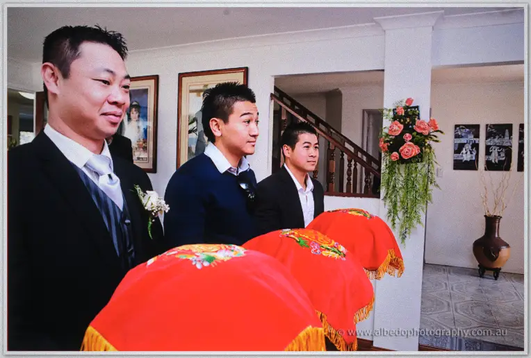 Groomsmen in dark suits stand behind red ceremonial gift trays in a bright home ahead of the tea ceremony.