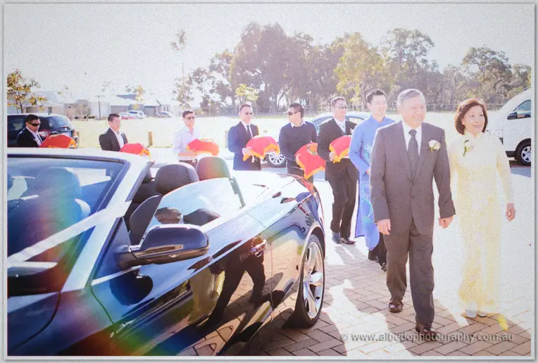 Khoa's party follows a ribboned convertible as his parents in ao dai lead the procession on a sunlit street.