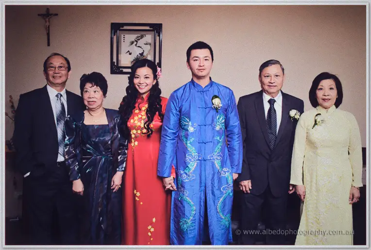 Michelle and Khoa in red and blue ao dai pose with both sets of parents beneath a crucifix at home.