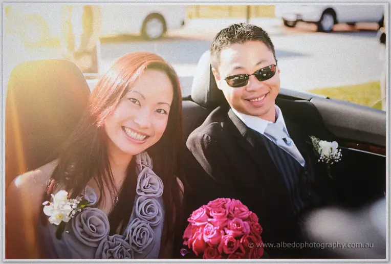 A bridesmaid rides the open-top wedding car in sunglasses, grinning as sunlight flares across the street.