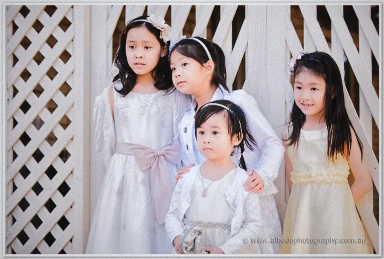 Four little flower girls in white and cream dresses huddle by a lattice gate, the shy youngest held close.