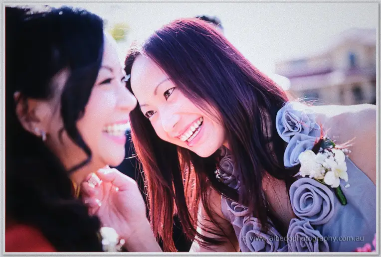 The bride and her maid of honour share a laughing moment outdoors, forehead to forehead in soft backlight.