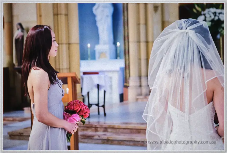 Bridesmaid in grey with a red rose bouquet waits at the front as the veiled bride faces the sanctuary.