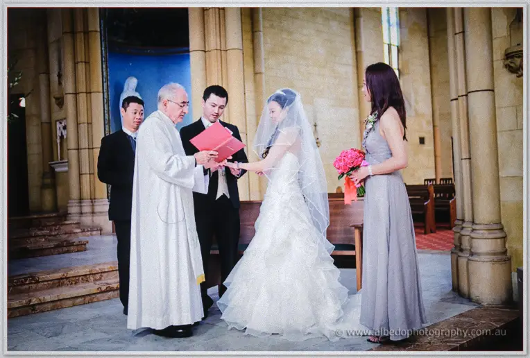 The priest blesses Michelle and Khoa's joined hands at the altar while a bridesmaid in grey looks on.