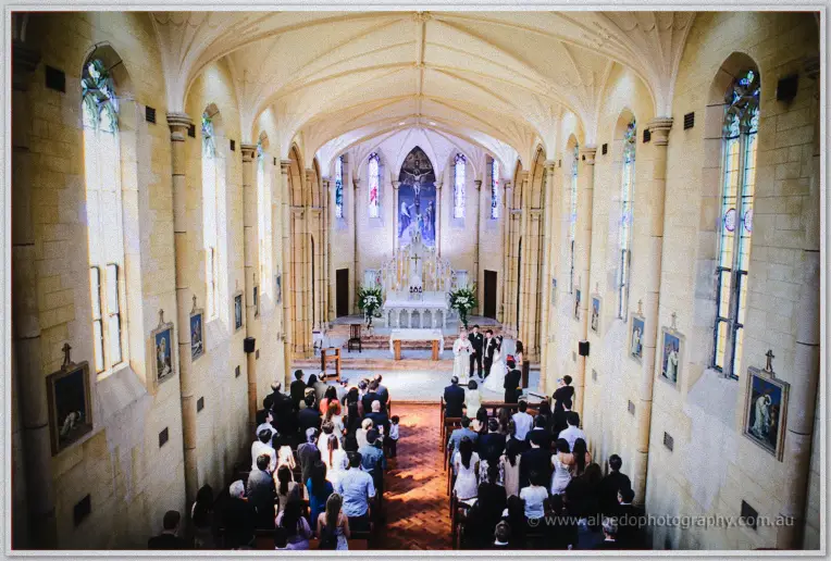 Wide view of the sandstone church as guests rise around the couple exchanging vows beneath the crucifix.