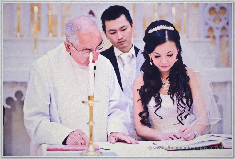 Michelle signs the marriage register in a tiara and veil as the priest and Khoa lean in close by candlelight.
