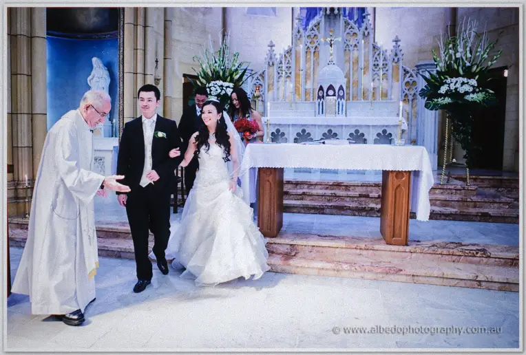 Newlyweds Michelle and Khoa walk arm in arm from the altar, the priest smiling as they pass.