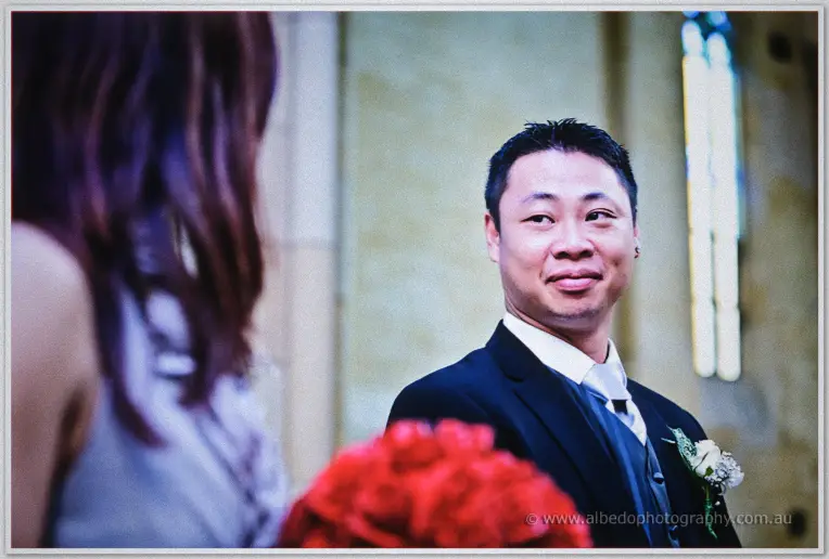 Close-up of Khoa's soft smile across the aisle, red roses blurred in the foreground of the church light.