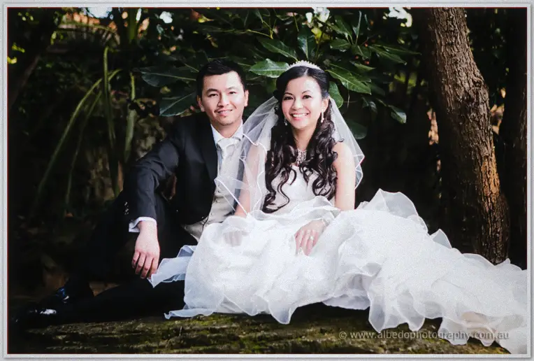 Seated on a mossy log under a broad tree, Michelle rests her hand on Khoa's chest.
