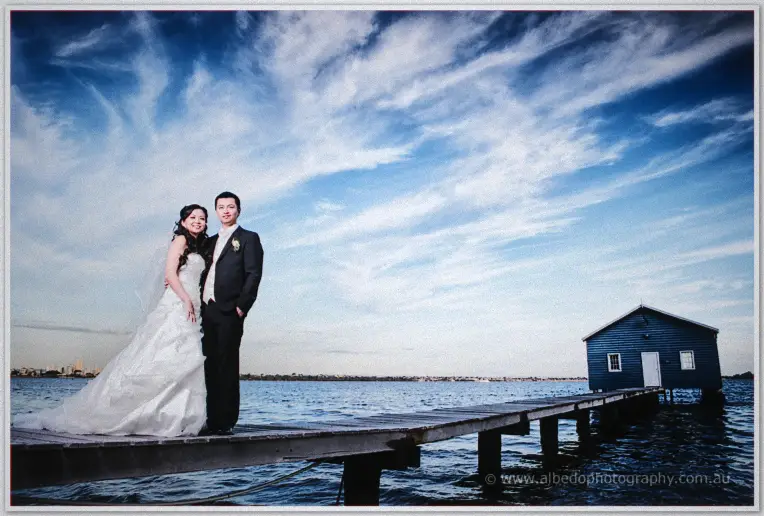 Michelle walks the timber jetty toward the blue boatshed, veil drifting over the calm water.