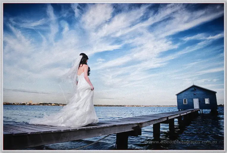Michelle stands mid-jetty gazing at the boatshed under a vast streaked sky.