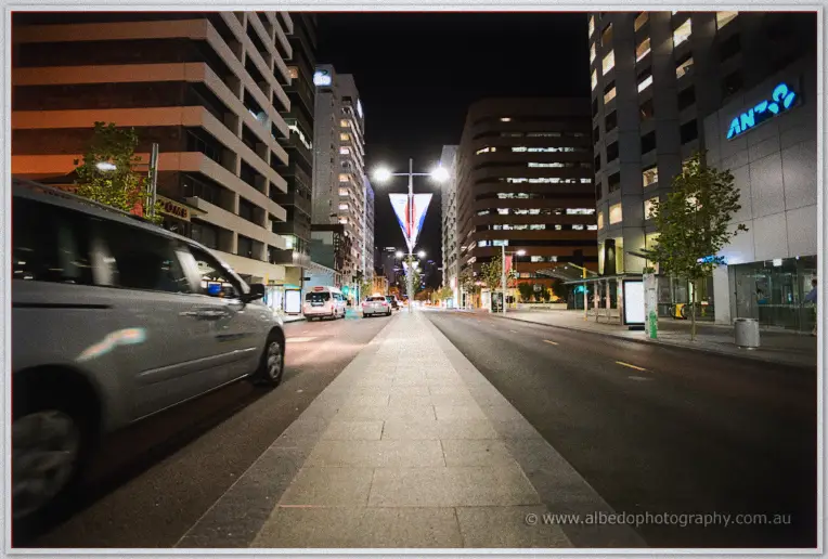 Long-exposure night view down a city street with passing cars and glowing office towers