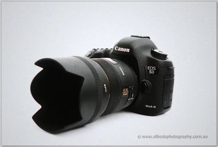 Black DSLR with a petal lens hood lies on a pale grey studio backdrop in a product-style still