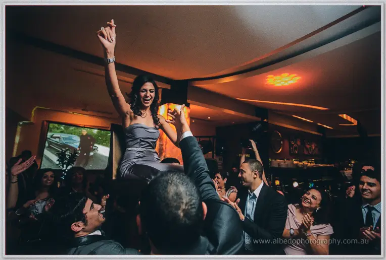 Cheering guests lift Lydia high on a chair, her fist punched into the party lights