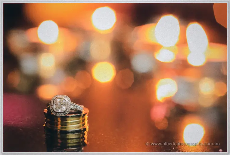 A diamond halo ring rests atop stacked gold bands against the blur of warm night lights