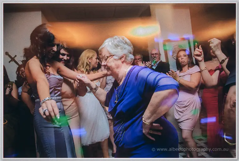 An elderly guest in royal blue dances joyfully with Lydia amid colour-flecked light