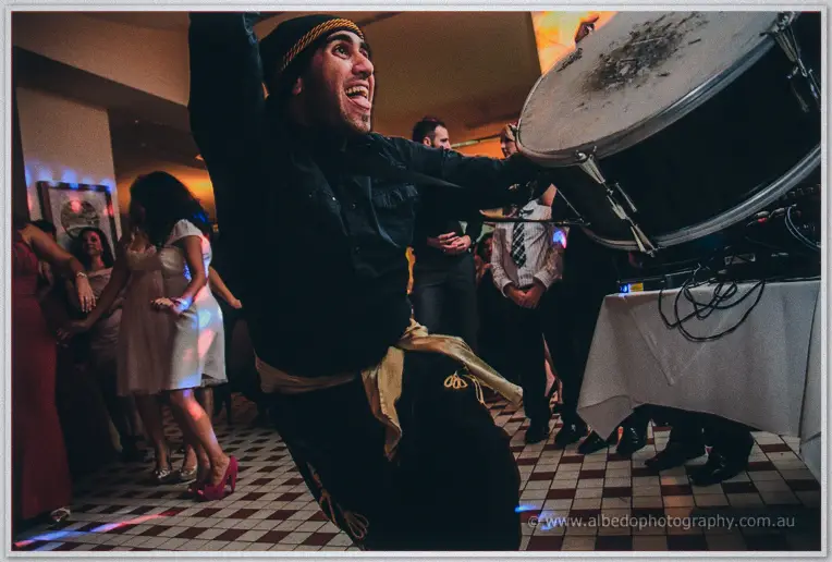 The drummer lunges mid-beat, head thrown back, gold sash flying across the chequered floor