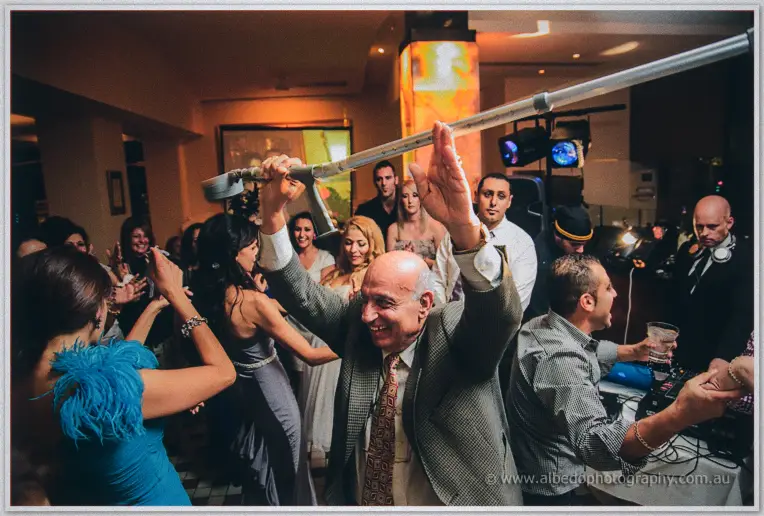 An elder waves his cane overhead as the blue-feathered guest dances past the wedding cake
