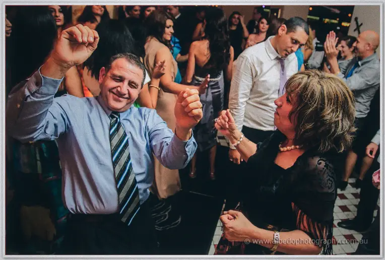 Wedding guests dance arm in arm under warm lights, the man mid-laugh in striped shirt and tie