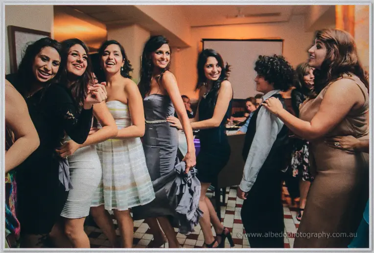 Women in cocktail dresses laugh and dance together on the checkered floor of the reception