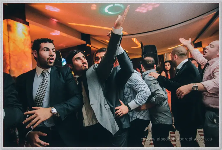 Men in suits dance with raised arms beside the speakers as colored lights sweep the party