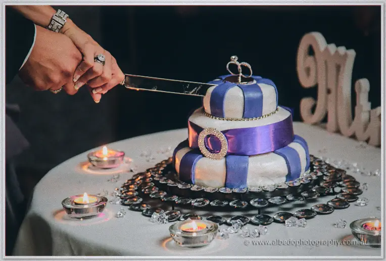 Hands clasped over a knife slicing the striped purple and white cake ringed by glowing tealights