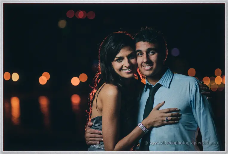 Lydia and Mina smile cheek to cheek after dark, city bokeh glinting on the water behind them
