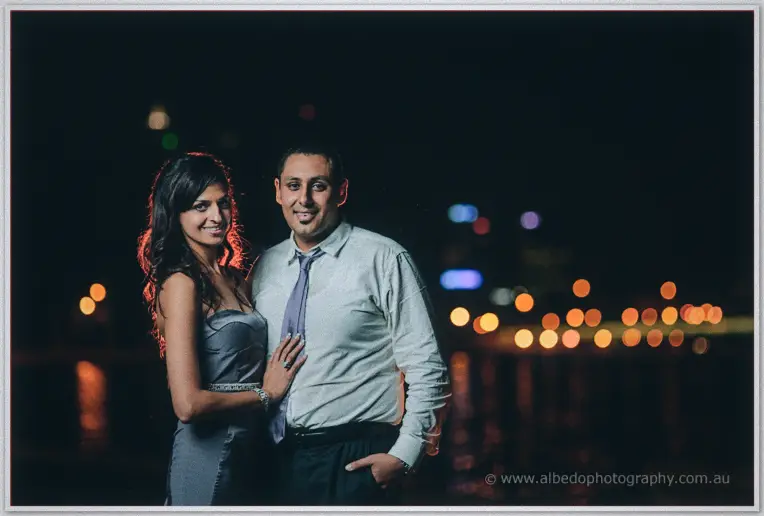 The couple stands close on the waterfront at night, city lights blurred across the dark water