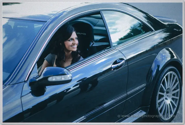 Lydia beams from the driver's seat of a polished black coupe in soft overcast light