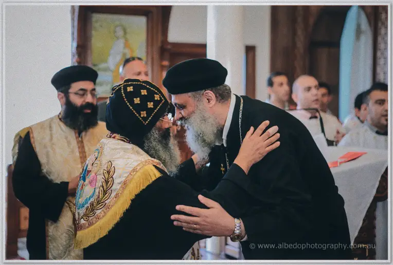 Two bearded Coptic priests embrace warmly at the altar as clergy and guests look on
