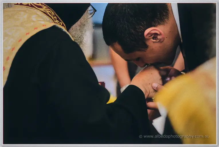 Mina bows to kiss the priest's hand in a close-up of reverence and tradition