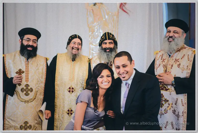 Lydia and Mina grin alongside four bearded priests in cream vestments after the ceremony